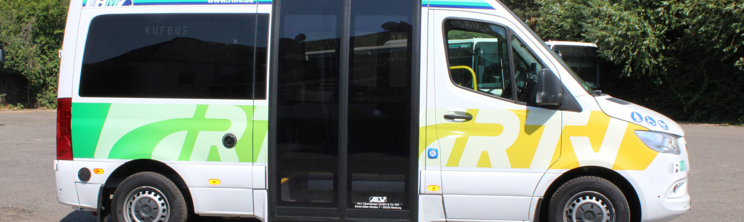 Seitliche Gesamtansicht eines modernen Rufbusses mit großem Schiebetürbereich, gelb-grüner RTV-Grafik und RMV-Logo. Auf der Tür sind Piktogramme für Barrierefreiheit, Kinderwagen und Fahrradmitnahme zu sehen.